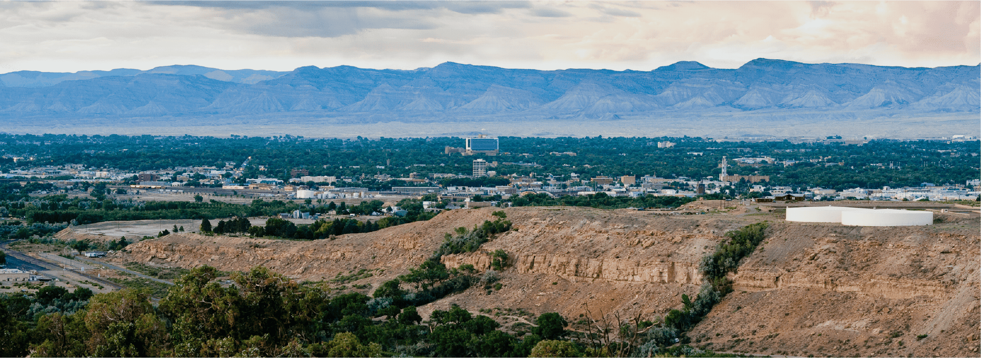 A panoramic view of a city surrounded by greenery, with rolling hills and tall mountains highlights the commitment to affordable health care, making it an ideal landscape for Montrose Health Care’s mission under a partly cloudy sky.