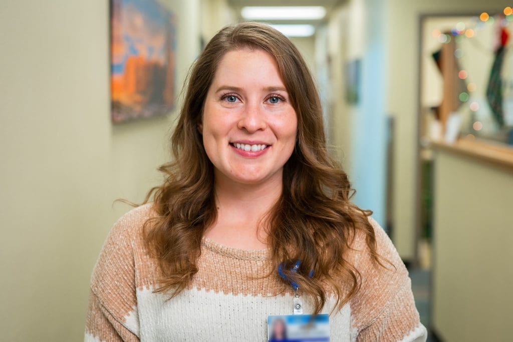 A woman with long brown hair wearing a beige and white sweater smiles at the camera in a brightly lit hallway at Montrose Health Care, with art on the walls and a decorated office area in the background.