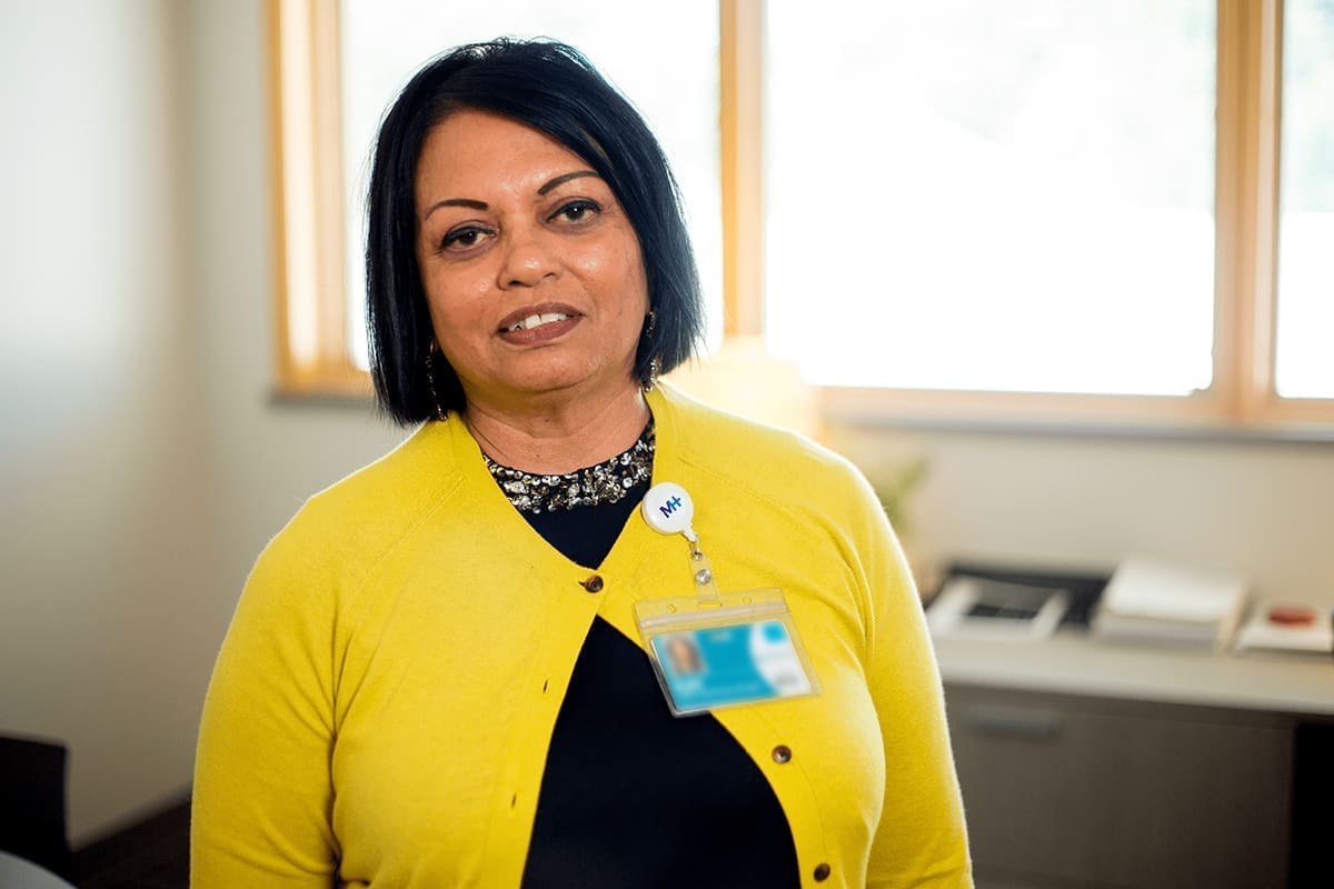 A woman with shoulder-length dark hair wearing a yellow cardigan and a Montrose Health Care name badge stands in an office with large windows and a desk in the background.