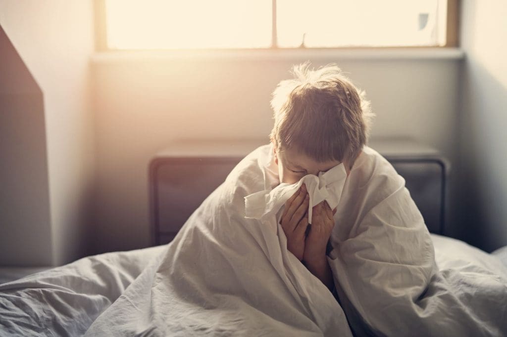 A young child wrapped in a white blanket sits on a bed, holding a tissue to their nose and appearing to sneeze, with sunlight streaming through a window—Montrose Health Care is here for moments like these.
