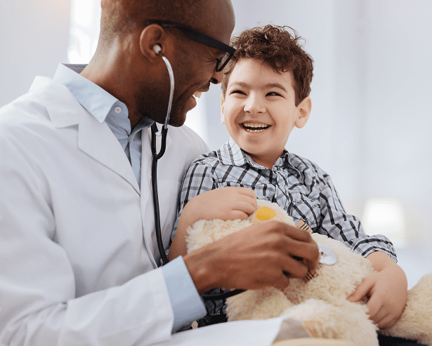A smiling doctor uses a stethoscope on a teddy bear held by a laughing young boy, both sharing a joyful moment during a Montrose Health Care checkup in a bright, friendly office.