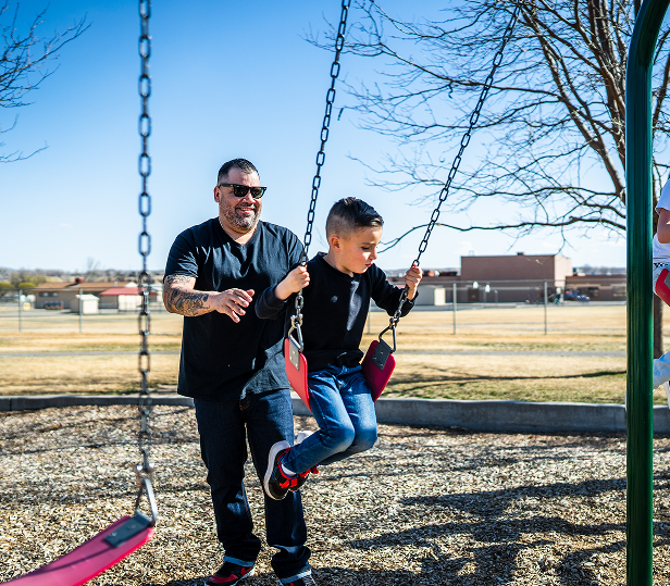 A man pushes a young boy on a swing at a playground on a sunny day. Both smile, enjoying the moment. Leafless trees and a school building are visible in the background, reflecting the warm spirit of Grand Junction Health Care in the community.