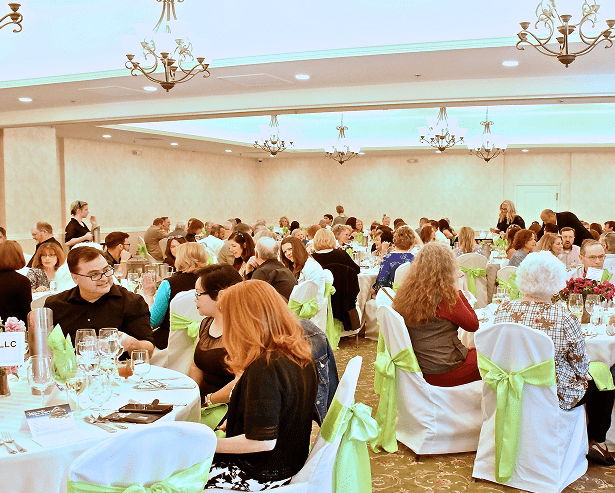 A large group of people sit at round tables covered with white tablecloths and green chair sashes in a banquet hall, dining and socializing under chandeliers at a Montrose Health Care event supporting affordable health care.