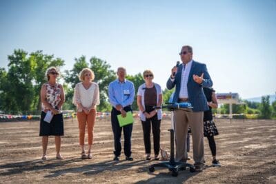 Six adults stand outdoors on a dirt lot, with one man in a suit speaking into a microphone at a podium about Affordable Health Care. The others listen attentively. Trees and a clear blue sky are in the background.