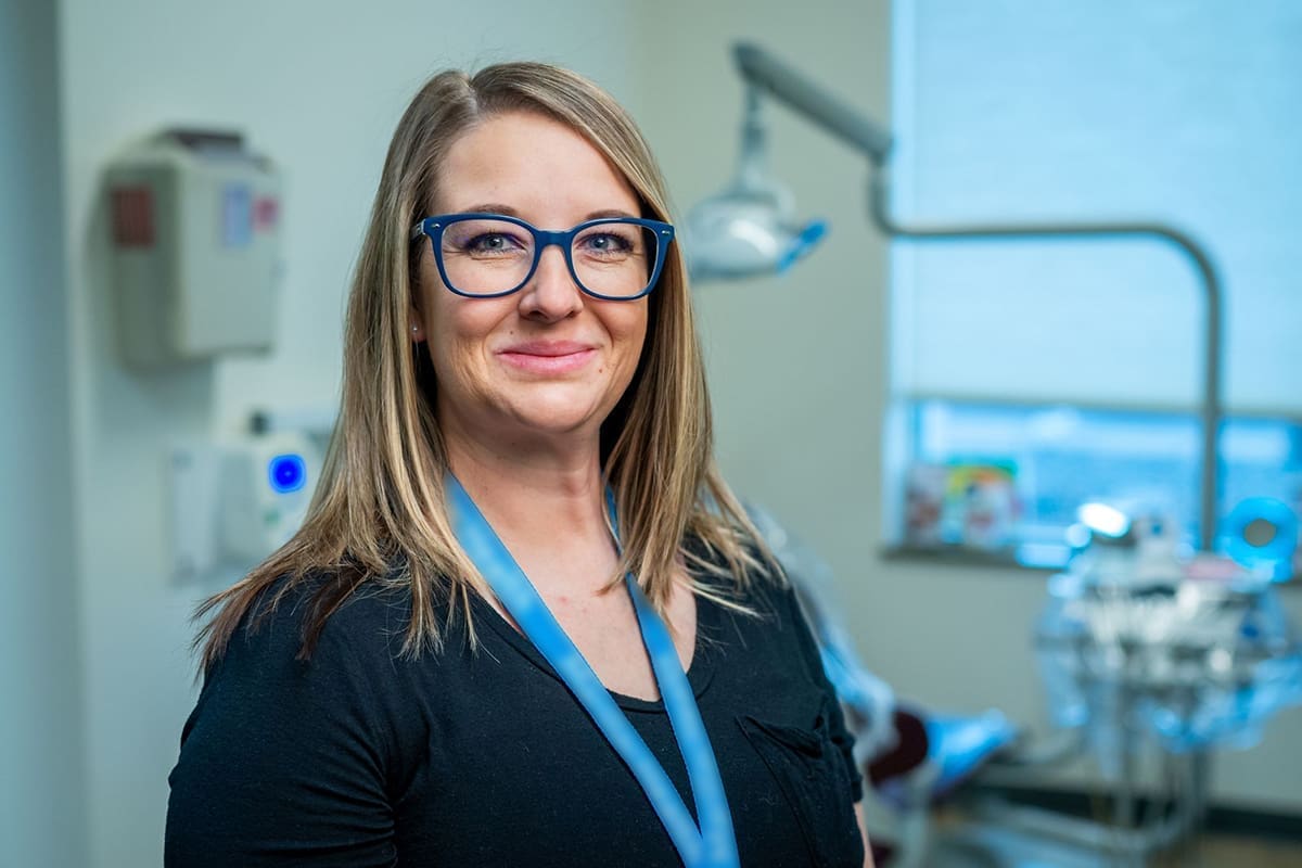 A woman with long blonde hair and blue glasses smiles while standing in a Grand Junction Health Care dental office, wearing a black top and blue lanyard. Dental equipment and a window are visible in the background.