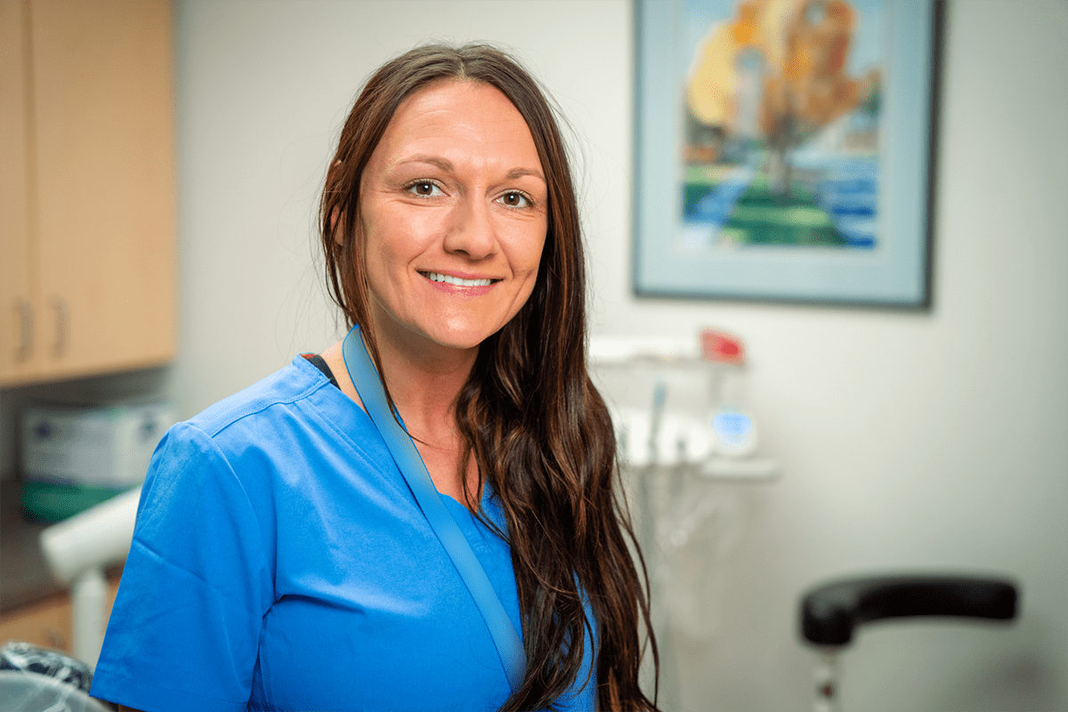 A woman in blue medical scrubs smiles while standing in a brightly lit Grand Junction Health Care clinic room with dental equipment and artwork in the background.
