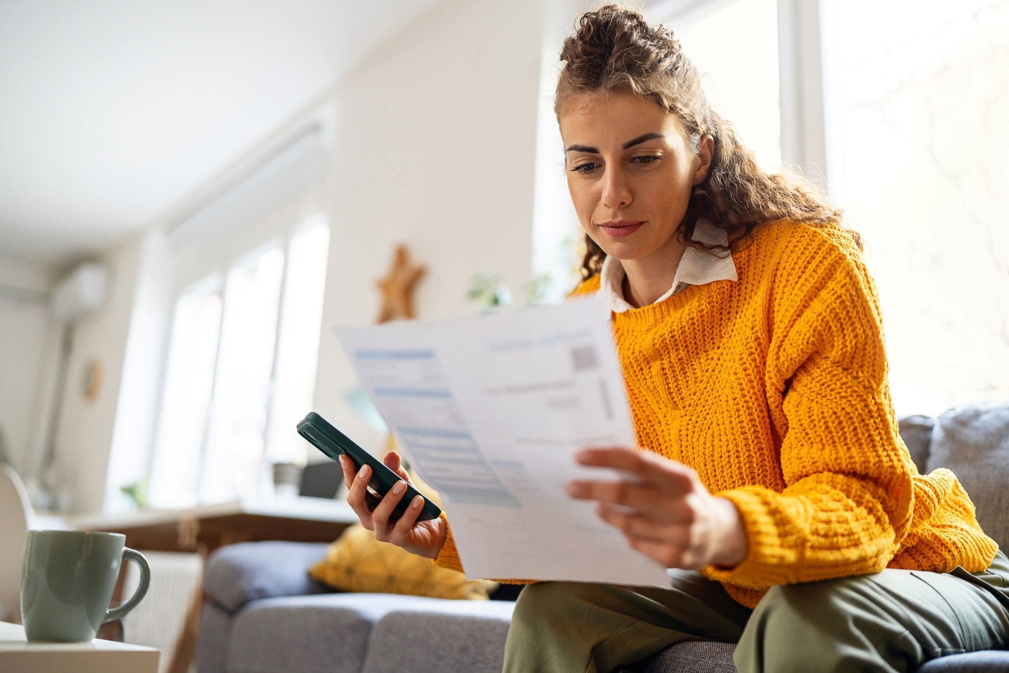 A woman in a yellow sweater sits on a couch, holding a smartphone and reading an Affordable Health Care document. A mug and papers are on the table in front of her. The setting is a bright, cozy living room.