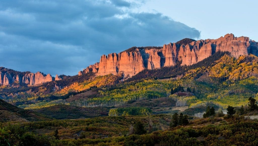 Dramatic red rock cliffs are illuminated by sunset light above rolling hills covered in green and yellow trees, reflecting the tranquil beauty surrounding Montrose Health Care under a partly cloudy sky.