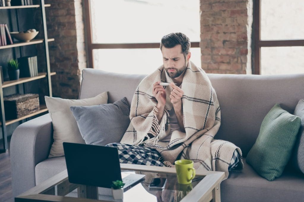 A man sitting on a couch wrapped in a plaid blanket looks at a thermometer with concern. He is wearing pajamas, with a laptop open on the table in front of him, searching for affordable health care in his cozy living room.