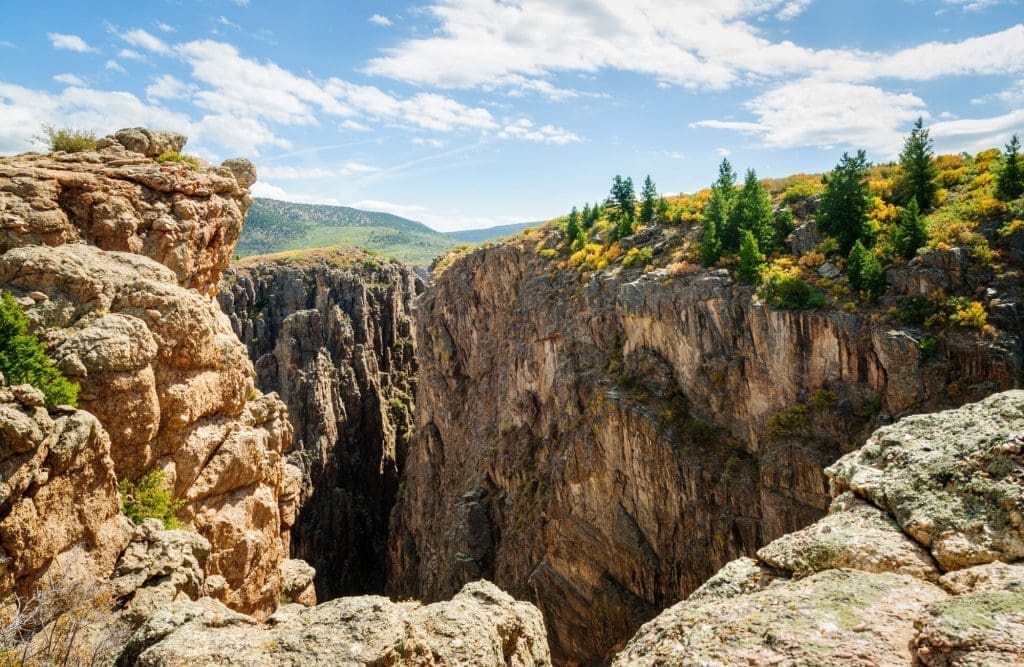 A deep, rocky canyon with steep cliffs and scattered trees on top, under a partly cloudy blue sky. Green hills and more trees are visible in the background, symbolizing the pursuit of affordable health care amidst nature’s grandeur.