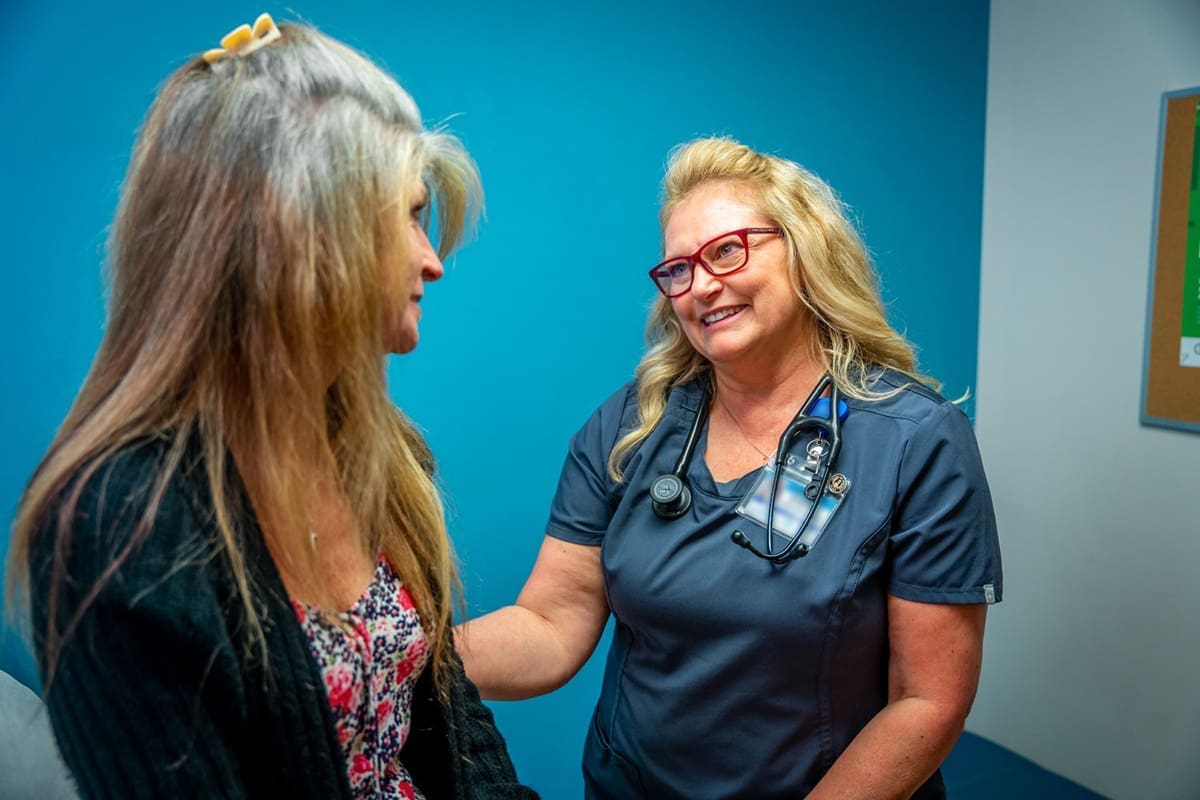 A Montrose Health Care professional with a stethoscope smiles and talks to a seated patient with long hair in a clinical room with blue walls, reflecting the commitment to affordable health care.